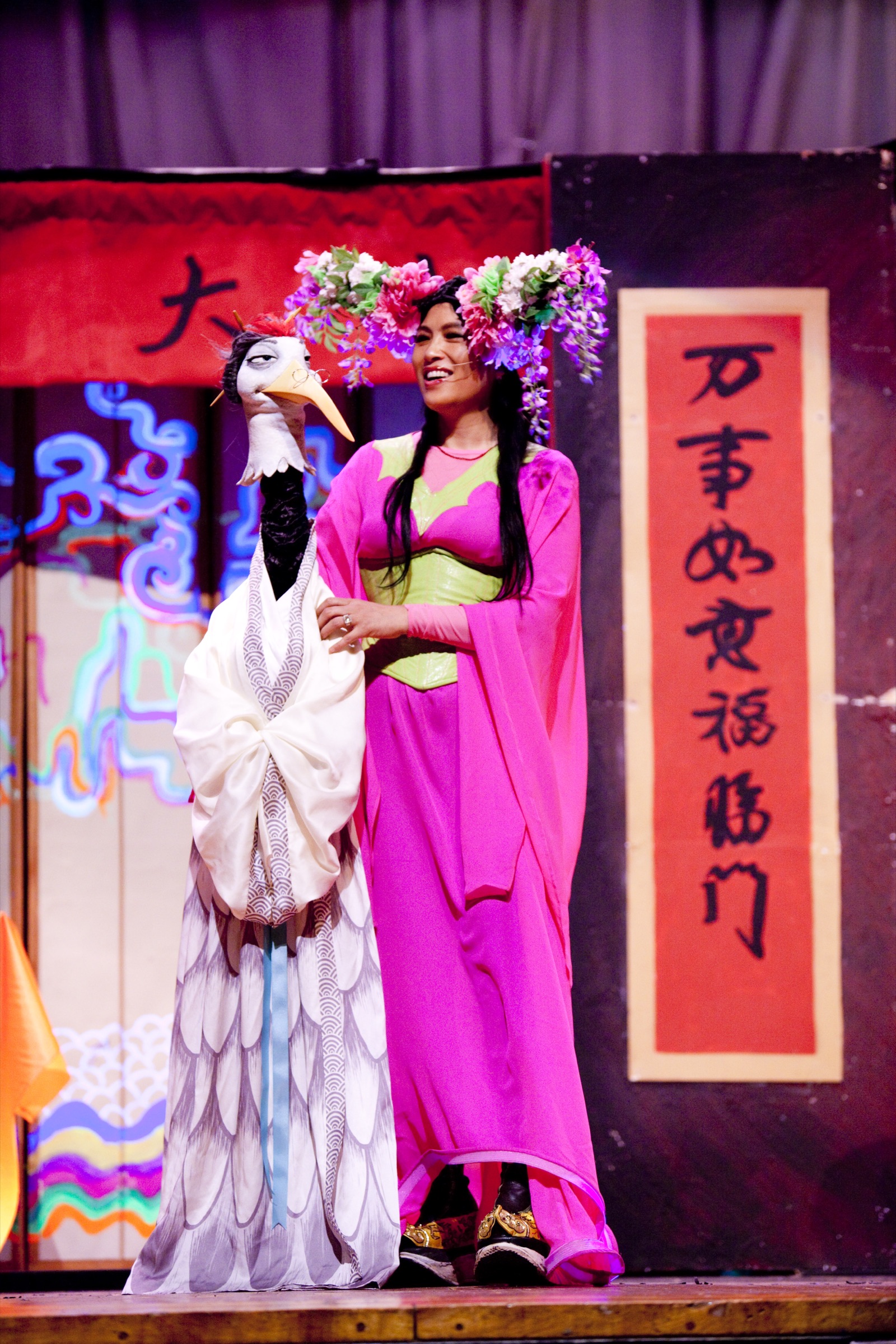 Grace Chang in flower crown and pink robes beside a white crane puppet
